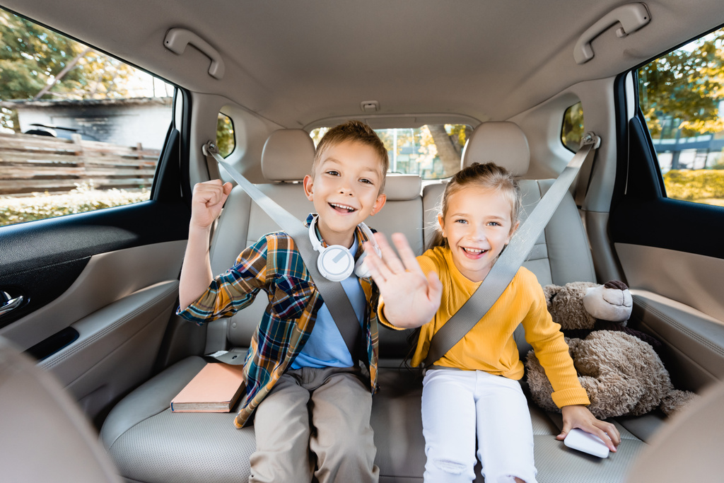 Χαμογελώντας παιδιά δείχνουν ναι και χαιρετώντας κάμερα κοντά smartphone και βιβλίο στο αυτοκίνητο  - Φωτογραφία, εικόνα