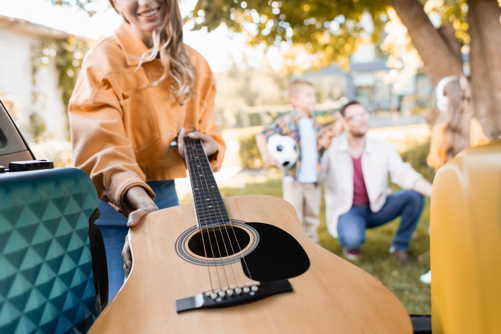 Vista recortada de la mujer sonriente tomando la guitarra acústica del maletero del coche cerca de la familia sobre fondo borroso al aire libre  - Foto, imagen