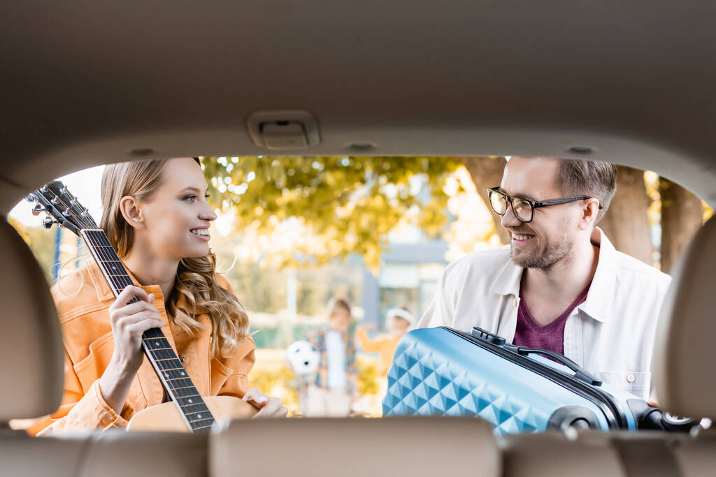 Wesoła para patrząca na siebie trzymając gitarę akustyczną i walizkę w pobliżu samochodu na niewyraźnym pierwszym planie  - Zdjęcie, obraz