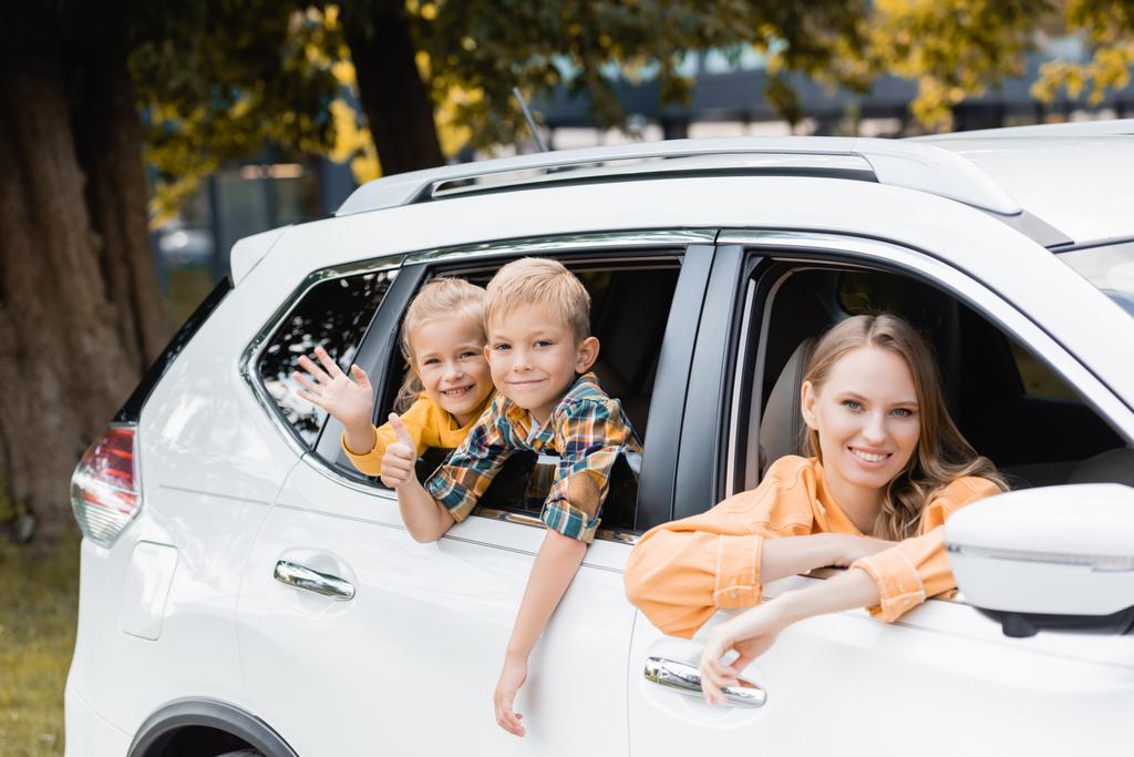Улыбающиеся дети машут и показывают, как рядом с матерью в машине во время отпуска  - Фото, изображение