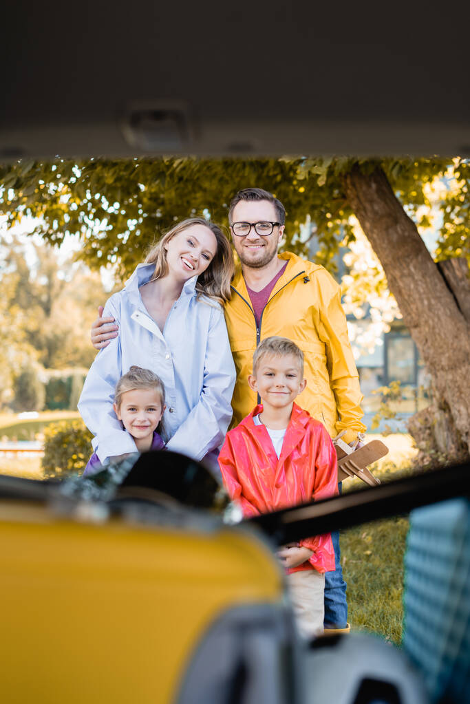 Lächelnde Familie blickt auf verschwommenem Vordergrund in die Kamera am Kofferraum des Autos  - Foto, Bild