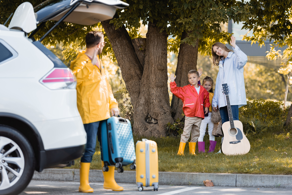 Familia en traje de otoño con guitarra acústica saludando al padre con la maleta cerca del coche en primer plano borroso al aire libre  - Foto, imagen