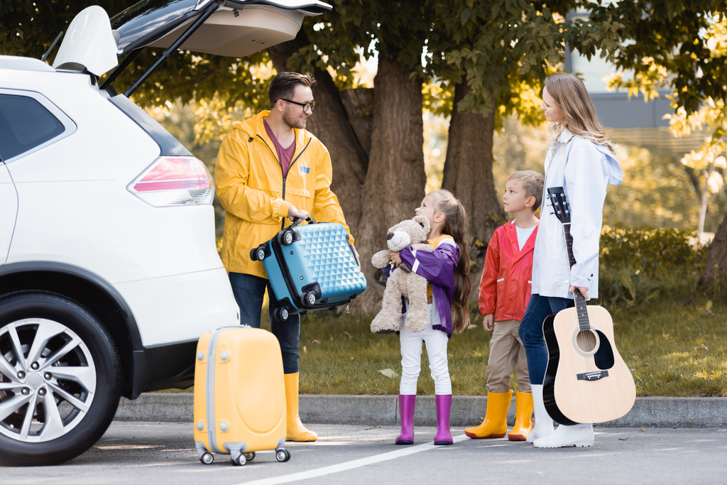 Uśmiechnięty mężczyzna w jesiennym stroju trzymając walizkę w pobliżu rodziny z zabawką i gitara akustyczna i auto na zewnątrz  - Zdjęcie, obraz