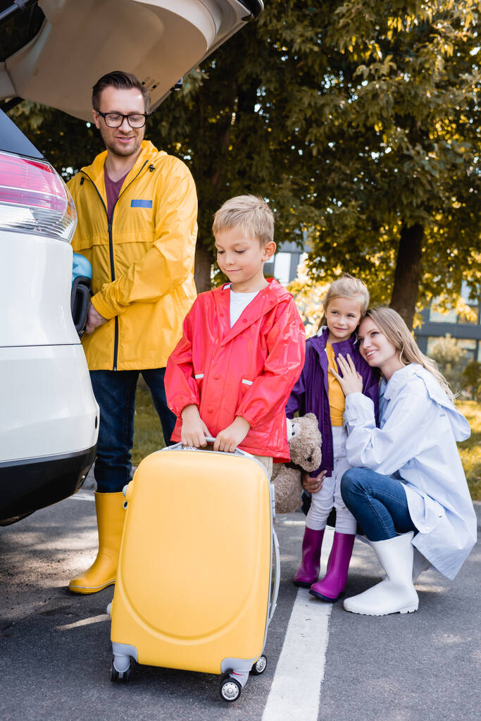 Uśmiechnięty chłopiec w jesiennym stroju trzyma walizkę w pobliżu rodziny i samochodu na zewnątrz  - Zdjęcie, obraz