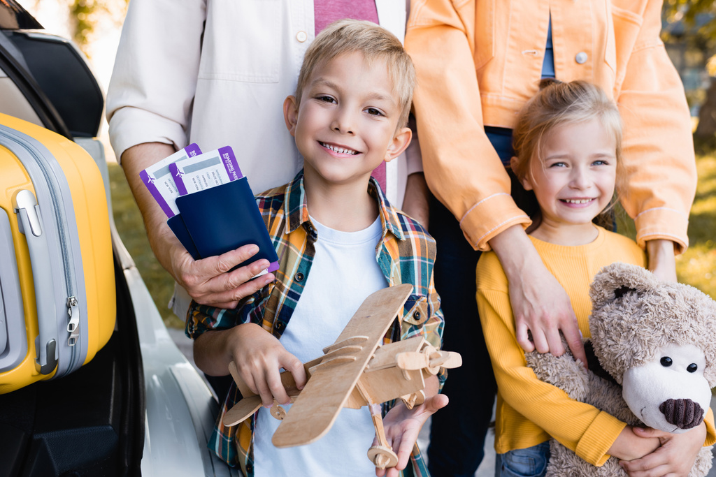 Lächelnde Kinder mit Spielzeug stehen neben Eltern mit Pässen und Koffer im Auto im Freien  - Foto, Bild