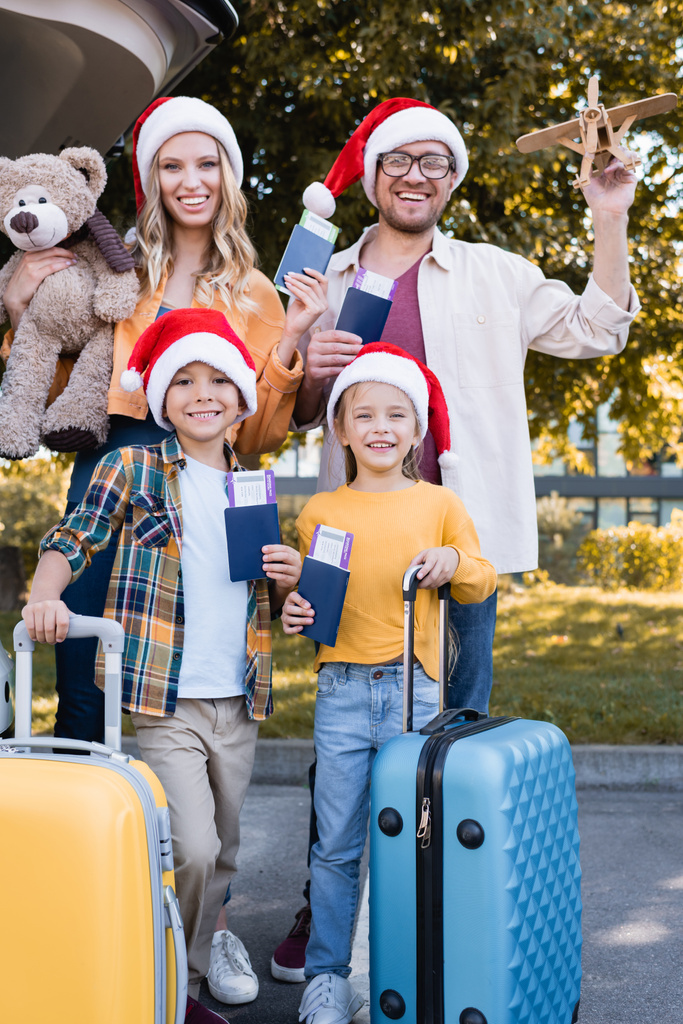 Wesoła rodzina w kapeluszach Mikołaja trzymająca paszporty w pobliżu walizek i samochodu na zewnątrz  - Zdjęcie, obraz