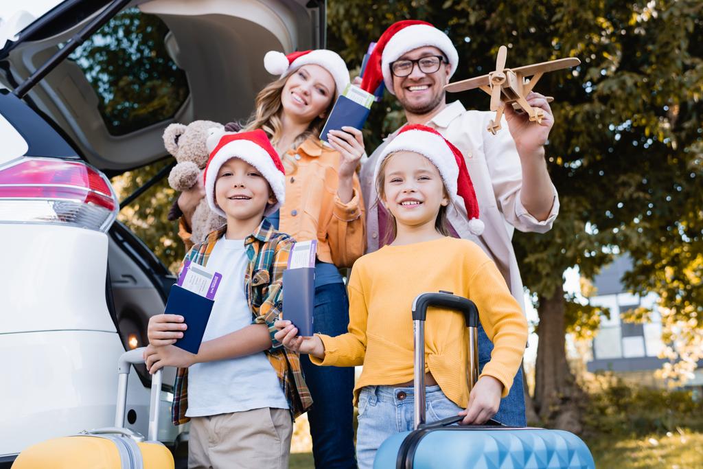 Familie in Weihnachtsmannmützen mit Pässen mit Flugtickets in der Nähe von Koffern und Autos im Freien  - Foto, Bild