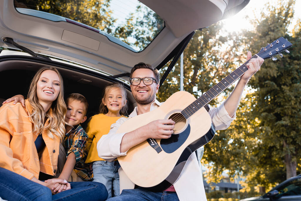 Χαρούμενη οικογένεια με ακουστική κιθάρα κοιτάζοντας κάμερα κοντά στο φορτηγό του αυτοκινήτου  - Φωτογραφία, εικόνα