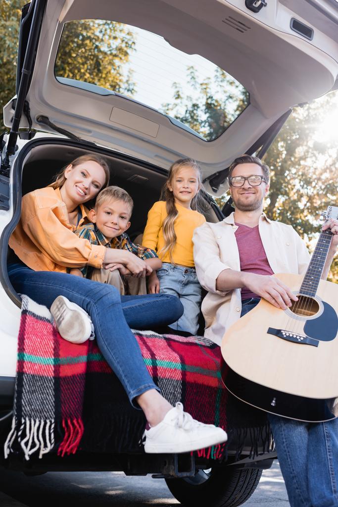 Улыбающиеся дети сидят рядом с родителями с акустической гитарой в багажнике автомобиля  - Фото, изображение
