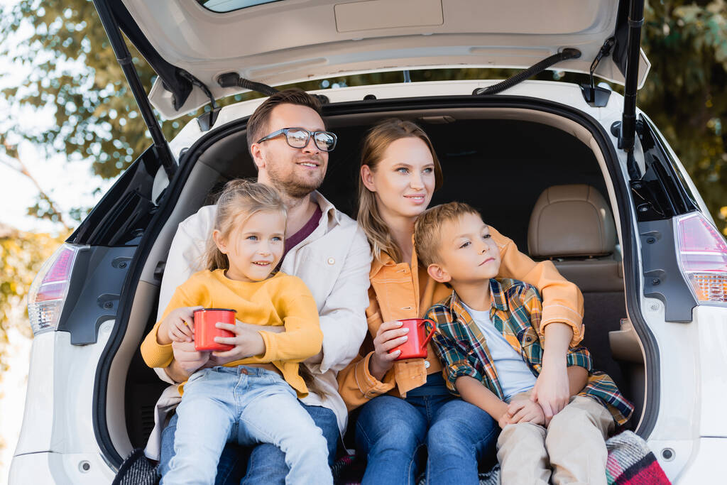 Parents souriants tenant des tasses et embrassant les enfants dans le coffre de la voiture pendant les vacances - Photo, image