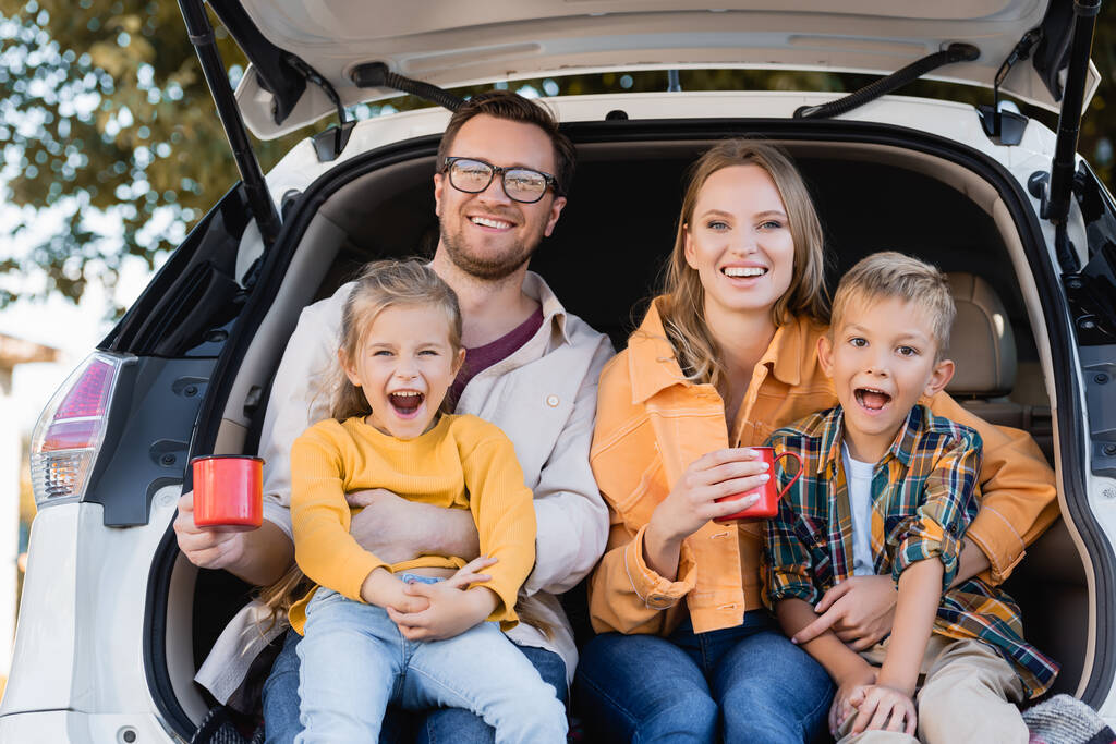 Vrolijke familie houden kopjes en kijken naar de camera terwijl zitten in de kofferbak van de auto  - Foto, afbeelding