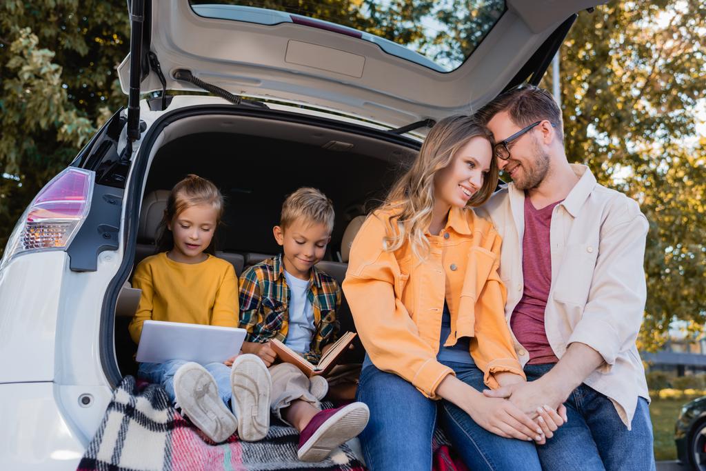 Bambini sorridenti con libro e tablet digitale seduti in camion di auto vicino ai genitori che si tengono per mano all'aperto  - Foto, immagini