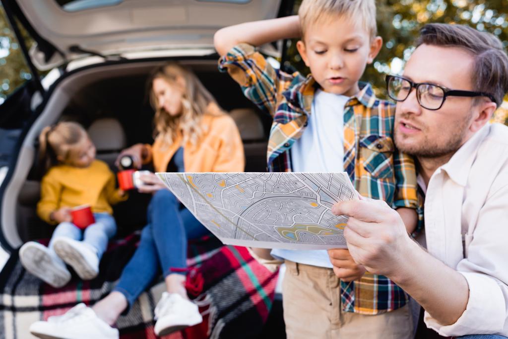 Padre sosteniendo mapa cerca pronto y familia en el maletero de coche al aire libre  - Foto, Imagen