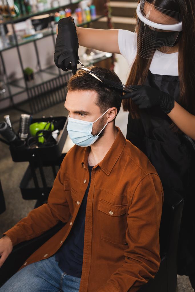 парикмахер в защитных и латексных перчатках стрижет волосы человека в медицинской маске - Фото, изображение