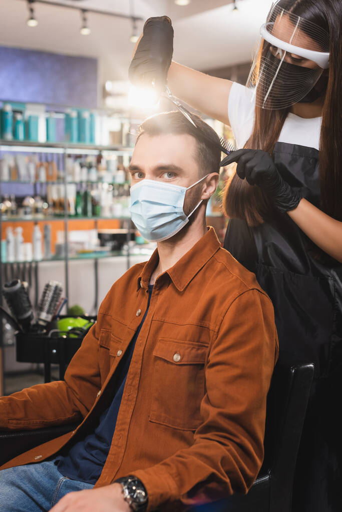 Mies lääketieteellisessä naamiossa katselee kameraa, kun kampaaja suojavarusteissa leikkaa hiuksensa - Valokuva, kuva