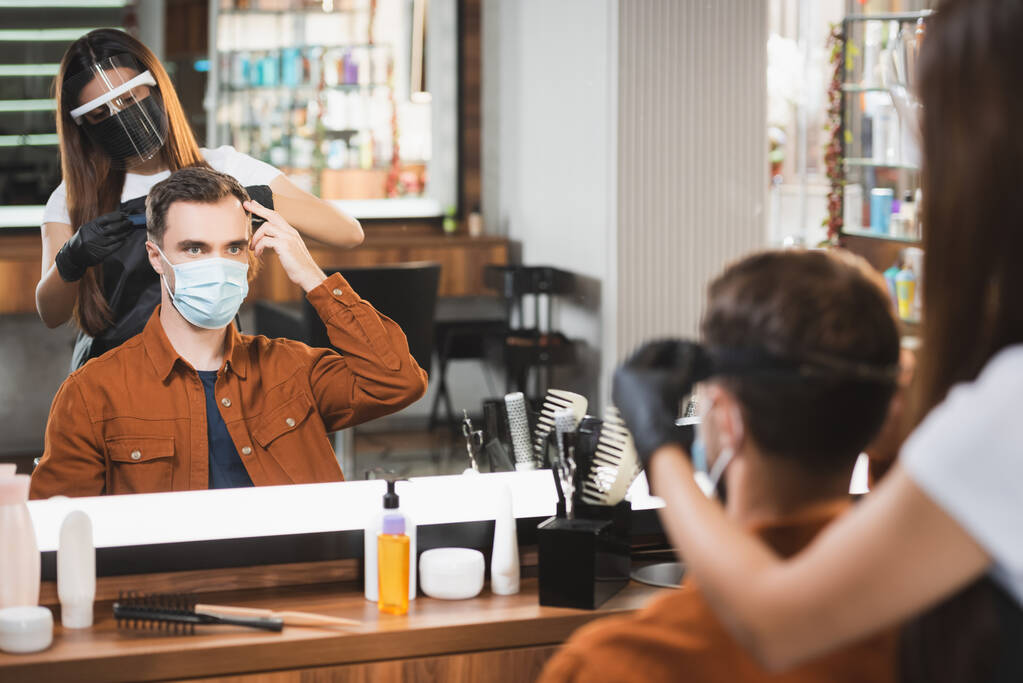 mirror reflection of hairdresser in face shield cutting hair of man pointing with fingers, blurred foreground - Photo, Image