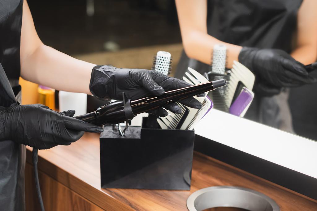 背景のぼやけた職場でカール鉄を保持するラテックス手袋の美容師のクロップドビュー - 写真・画像