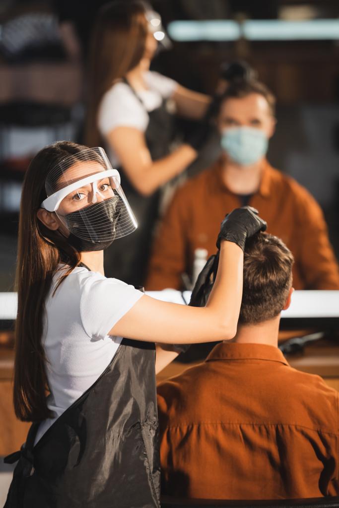 hairdresser in face shield looking at camera while touching hair of client on blurred background - Photo, Image