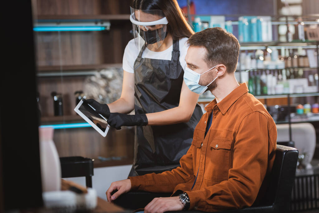 kapper in het gezicht schild en latex handschoenen met digitale tablet in de buurt van een man met medisch masker, wazig voorgrond - Foto, afbeelding