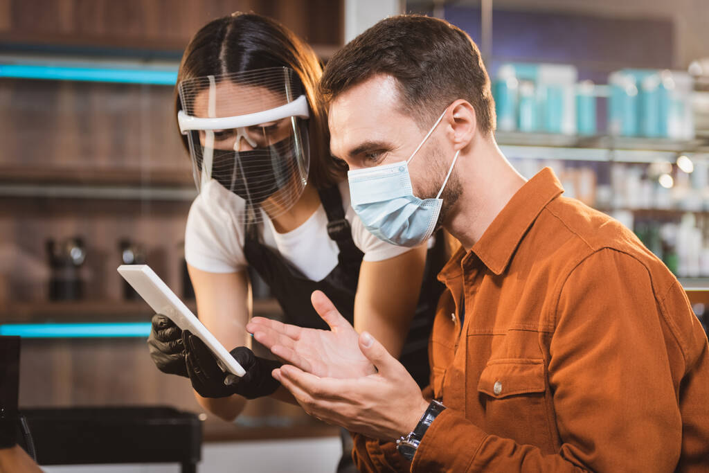 fryzjer w osłonie twarzy trzymając cyfrową tabletkę w pobliżu klienta w masce medycznej wskazując rękami - Zdjęcie, obraz
