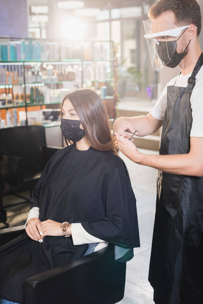 hairstylist in apron and face shield cutting hair of woman in medical mask - Photo, Image