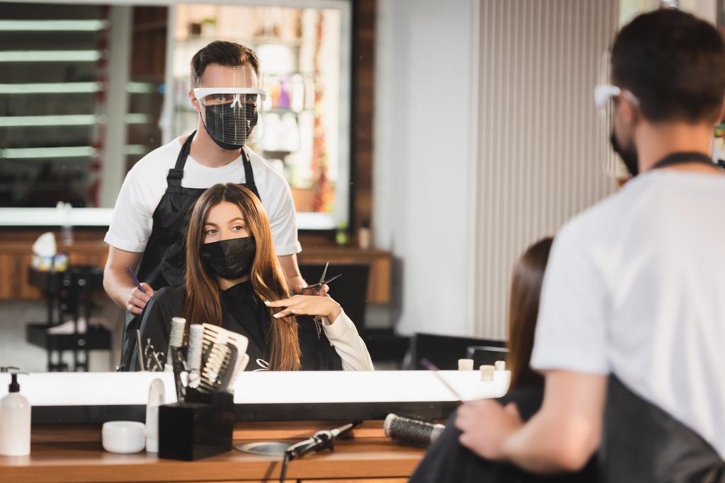 Spiegelreflexion des Friseurs in Schutzausrüstung in der Nähe des Kunden in medizinischer Maske, unscharfer Vordergrund - Foto, Bild
