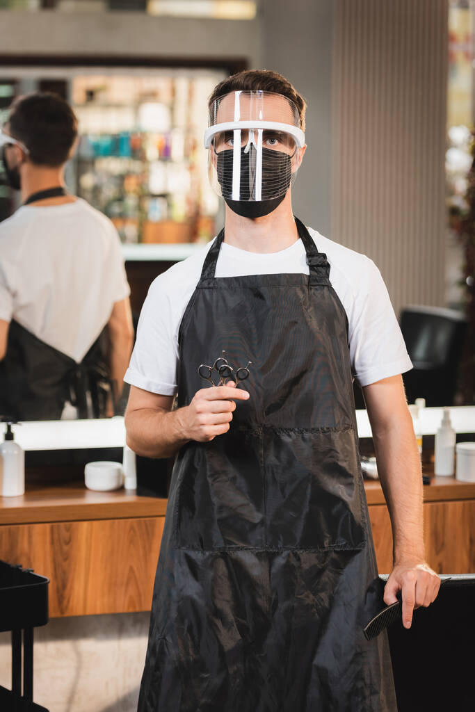 kadeřník v obličejovém štítu a zástěra drží nůžky při pohledu na fotoaparát na rozmazaném pozadí - Fotografie, Obrázek