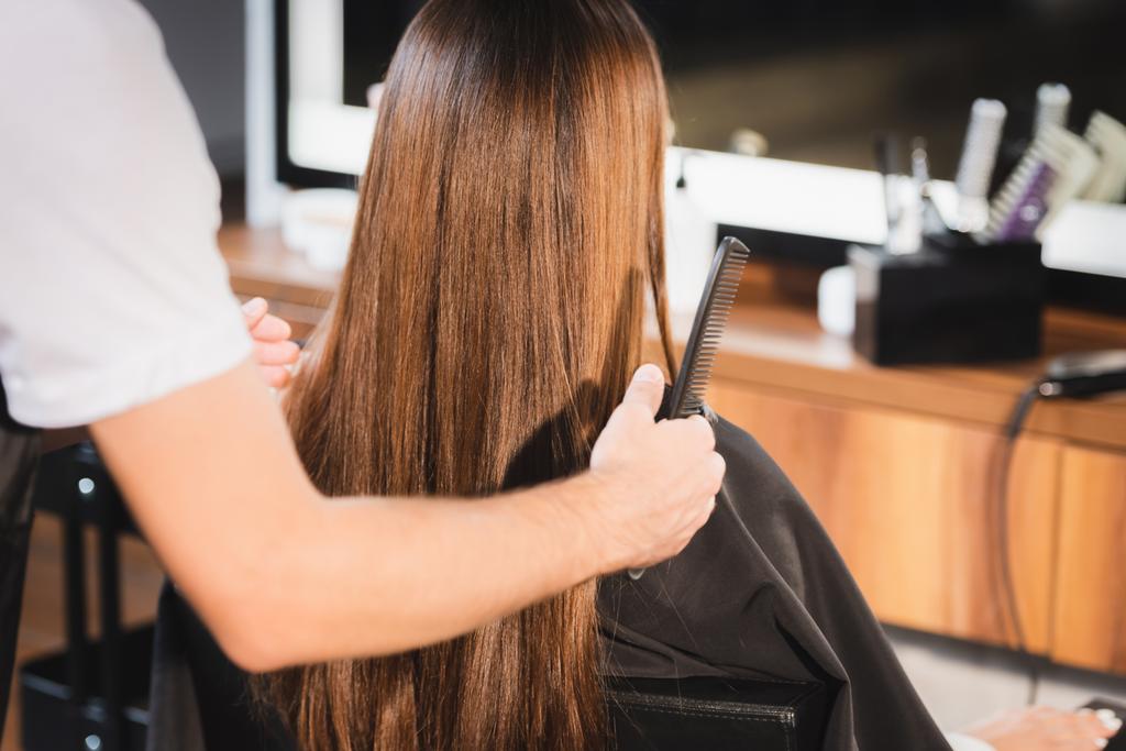 kadeřnice držící hřeben a dotýkající se vlasů ženy na rozmazaném popředí - Fotografie, Obrázek