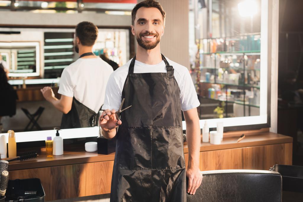 peluquero sonriente en delantal sosteniendo tijeras mientras está de pie en el lugar de trabajo sobre fondo borroso - Foto, imagen