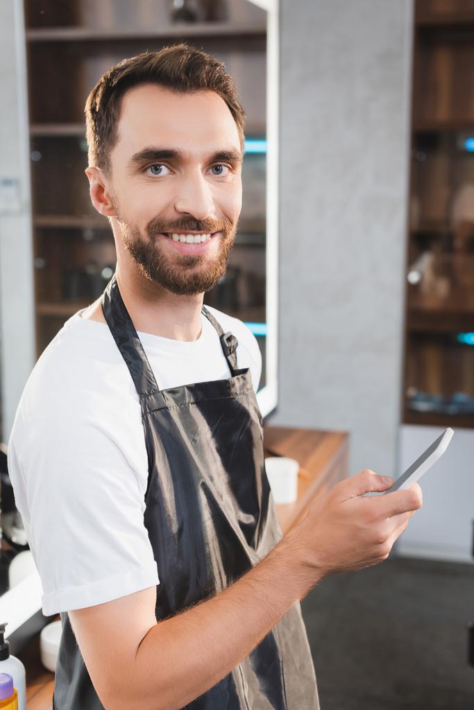 веселый, бородатый парикмахер смотрит в камеру во время чата на смартфоне - Фото, изображение