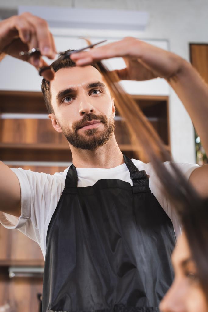 mladý vousatý kadeřník stříhání vlasů ženy na rozmazané popředí - Fotografie, Obrázek