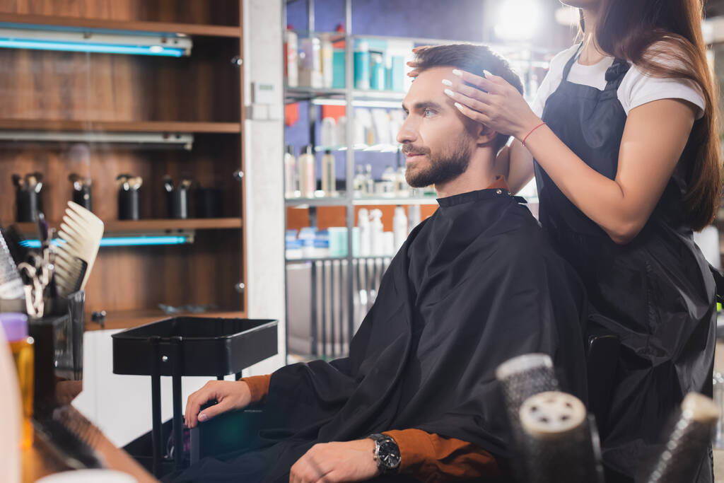 fryzjer dotykający włosów młodego brodatego mężczyzny, zamazany pierwszy plan - Zdjęcie, obraz