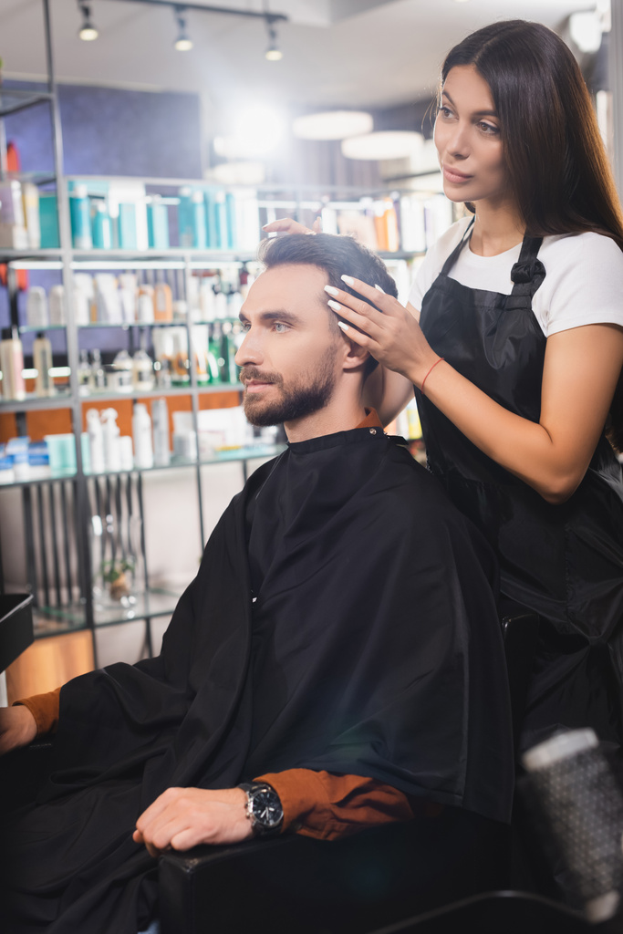 barber in apron touching hair of young bearded man in barbershop, blurred foreground - Photo, Image