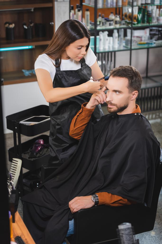 молодий перукар в фартусі, що ріже волосся людини, вказуючи пальцем
 - Фото, зображення