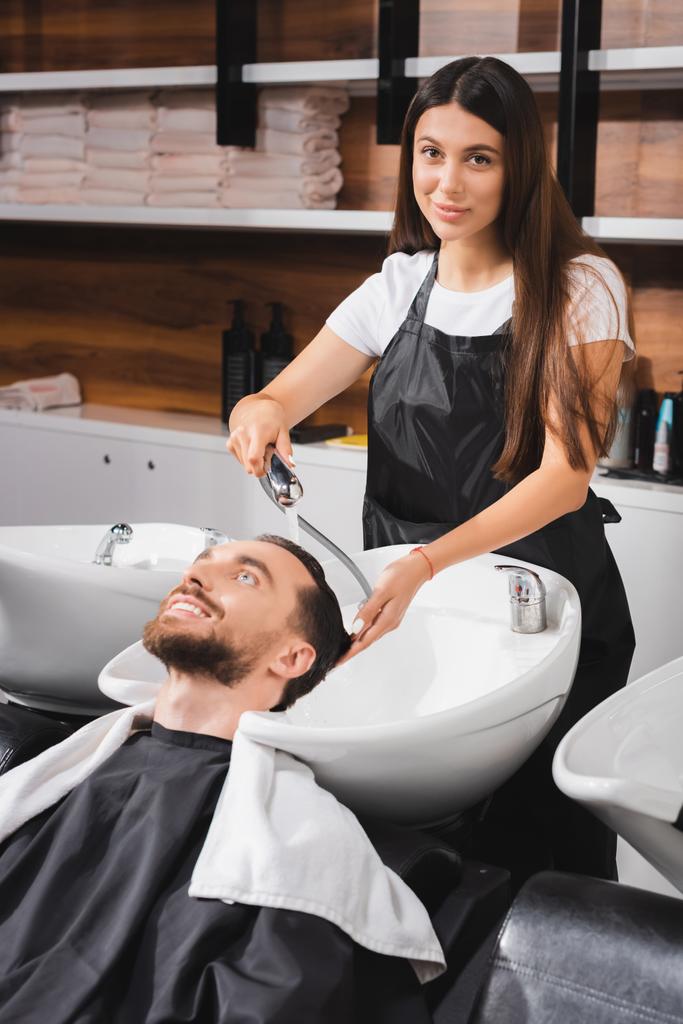 молодой парикмахер в фартуке, смотрящий в камеру во время мытья волос счастливого клиента - Фото, изображение