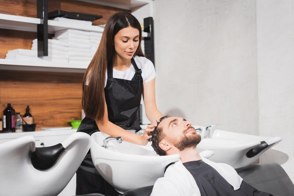 young hairstylist washing hair of bearded man in beauty salon - Photo, Image