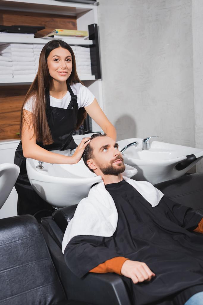 улыбающийся парикмахер смотрит в камеру во время мытья волос молодого человека  - Фото, изображение