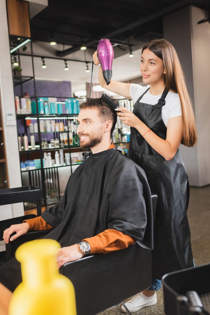 молодий перукар в фартусі сушить волосся усміхненого чоловіка, розмитий передній план
 - Фото, зображення