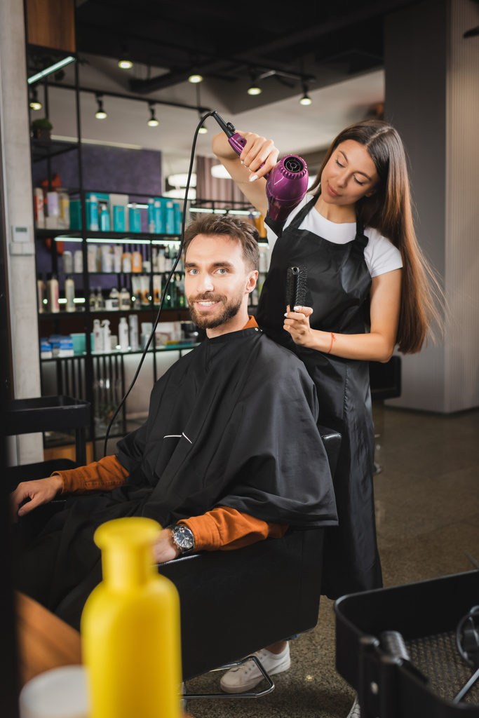 hombre barbudo feliz mirando a la cámara mientras la peluquería se seca el pelo, fondo borroso - Foto, Imagen