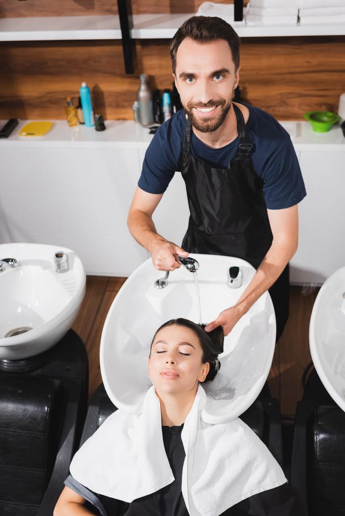 笑顔髭の美容師が理容室で若い女性の髪を洗う - 写真・画像