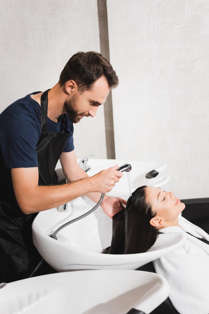 vue latérale du barbier barbu laver les cheveux de la jeune femme dans le salon de beauté - Photo, image