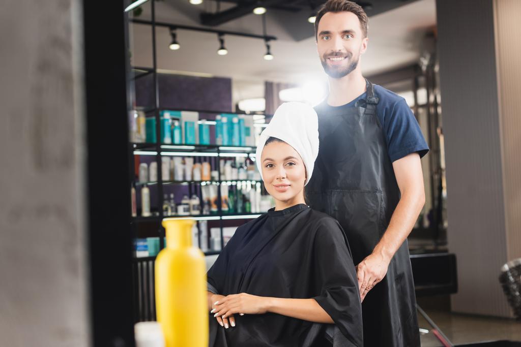 wesoły fryzjer i uśmiechnięta kobieta z włosami owiniętymi w ręcznik patrząc w kamerę, zamazany pierwszy plan - Zdjęcie, obraz