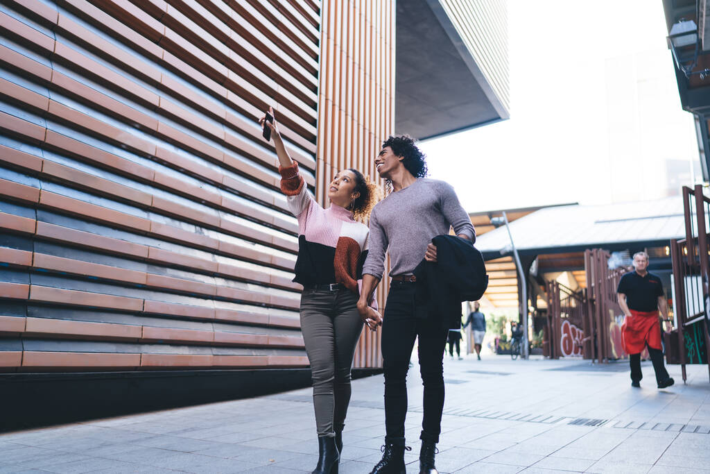 Heureux couple afro-américain en vêtements décontractés souriant et regardant loin tout en marchant dans la rue se tenant la main près du bâtiment moderne - Photo, image