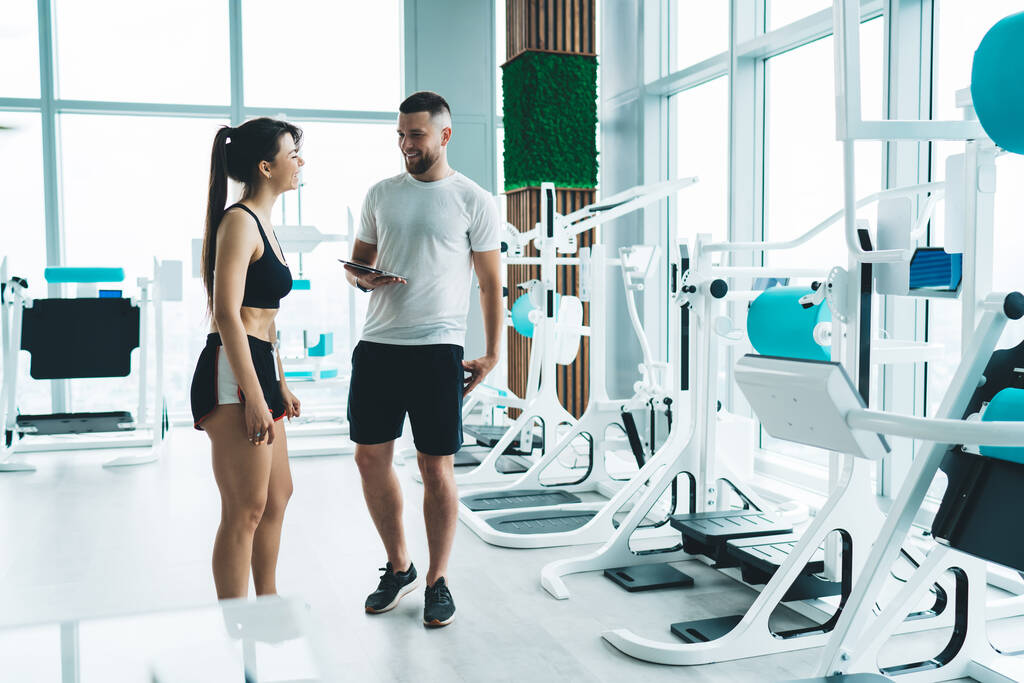 Περιεχόμενο νεαρή ταιριάζει θηλυκό σε activewear στέκεται με αρσενικό προσωπικό γυμναστή κρατώντας tablet σε στούντιο αστικής γυμναστικής κοντά σε εξοπλισμό γυμναστικής - Φωτογραφία, εικόνα