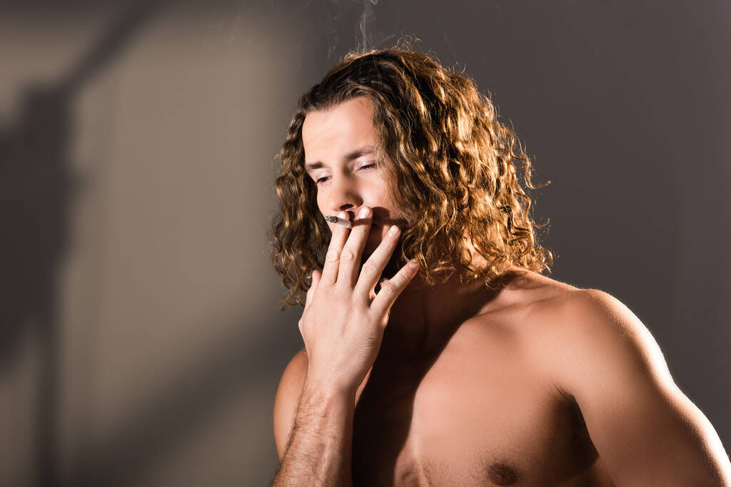 σέξι shirtless άντρας με μακριά μαλλιά καπνίζει σε σκούρο φόντο - Φωτογραφία, εικόνα