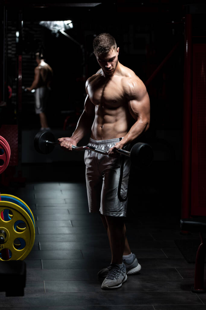 Мускулистый человек делает тяжелые упражнения веса для бицепсов с штангой в тренажерном зале - Фото, изображение