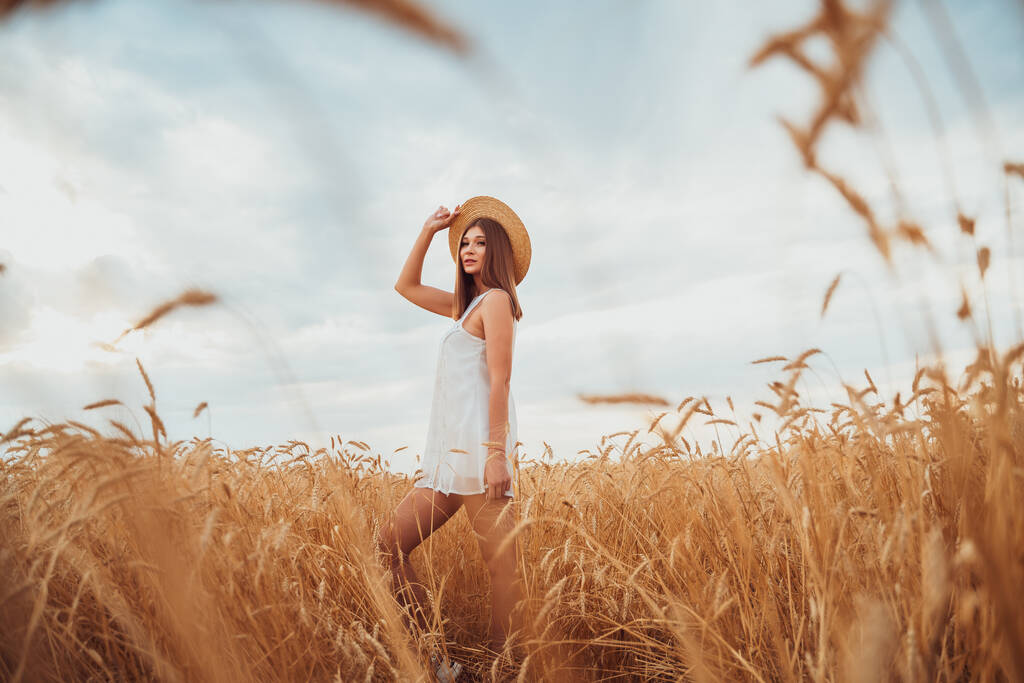 Giovane ragazza carina in un cappello di paglia e vestito bianco si trova in un campo di grano e gode di libertà - Foto, immagini