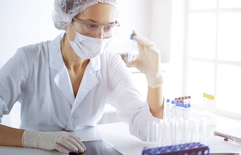 Professionele vrouwelijke wetenschapper in beschermende bril onderzoeken buis met reagentia in zonnig laboratorium. Geneeskunde en wetenschappelijk onderzoek - Foto, afbeelding