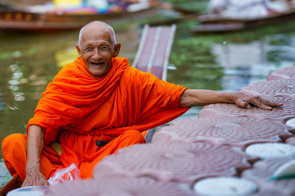 Buddhistischer Mönch sitzt in einem hölzernen Ruderboot im Kanal mit Almosenschale und wartet darauf, Essen von Dorfbewohnern rund um den schwimmenden Markt von Tha Kha in Samut Songkhram zu erhalten, einem berühmten Touristenziel in Thailand. - Foto, Bild