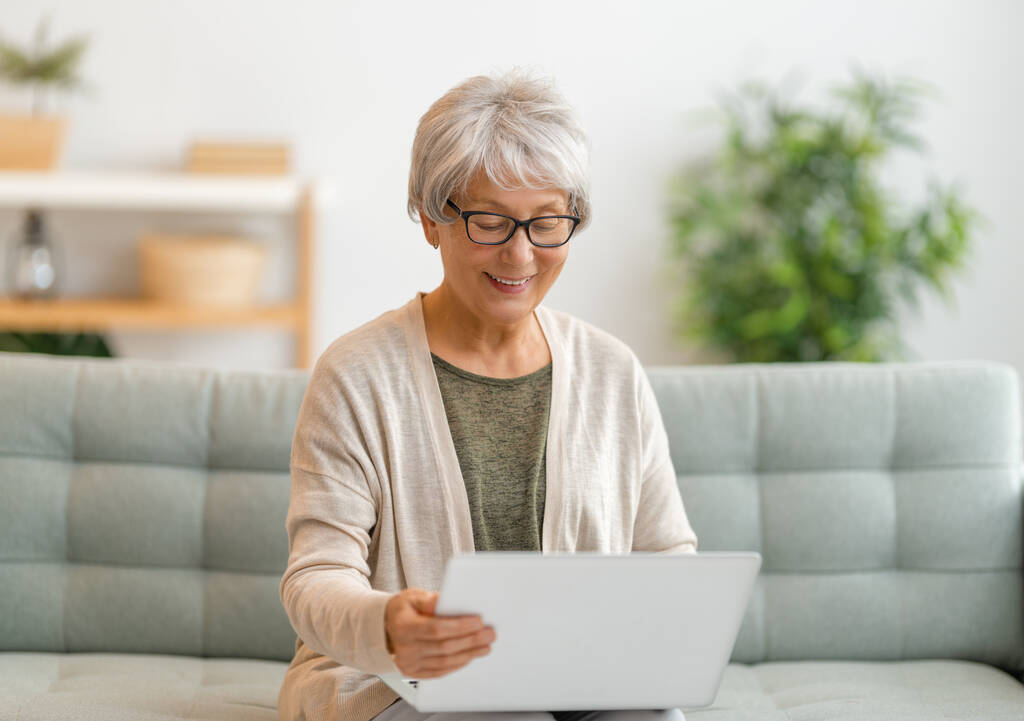 Radostná krásná starší žena používá notebook sedí na pohovce doma. - Fotografie, Obrázek